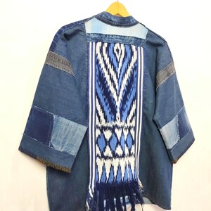 May include: A patchwork denim jacket featuring a central vertical panel of blue and white ikat fabric with a fringed hem. The jacket is constructed from various shades of blue denim fabric pieces, including light and dark blue sections, with three-quarter length sleeves and a collarless neckline.