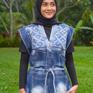 Puede incluir: Un chaleco de mezclilla sin mangas con cuello en V, cierre de un solo botón y cinturón para anudar. La prenda está confeccionada con paneles de mezclilla azul de diversos lavados y un patrón floral blanco estilo batik en el pecho.