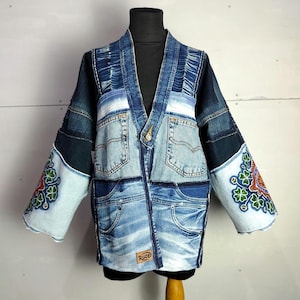 Könnte beinhalten: Eine Patchwork-Jeansjacke mit einzigartigem Design. Die Jacke weist verschiedene Blautöne auf, mit Taschen und dekorativen Nähten. Die Ärmel haben gestickte Blumenmuster in Rot, Grün und Orange. Die Jacke wird auf einer schwarzen Schaufensterpuppe präsentiert.