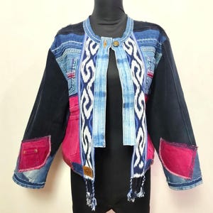 May include: A patchwork jacket constructed from blue denim, black fabric, and vibrant pink panels. The front features a central vertical band with a white and blue geometric pattern, a single button closure, and two dangling tassels at the hem.