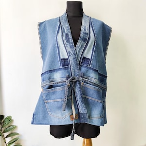 Pode incluir: Um colete de ganga sem mangas com um design de patchwork. O colete apresenta vários tons de ganga azul, bolsos e um fecho de laço. A peça de vestuário é exibida num manequim preto.