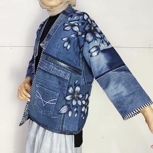 Könnte beinhalten: Jeansjacke mit floralem Design. Die Jacke ist ein Patchwork aus Jeansstoffteilen, mit bemalten weißen und grauen Blumen auf der Vorderseite und den Ärmeln. Die Jacke hat eine Tasche und einen schwarz-weiß gestreiften Besatz.