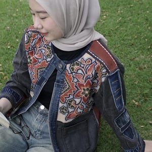 May include: A denim jacket with colourful floral patchwork on the upper back and sleeves. The jacket has a button-up front and multiple pockets. The person in the image is wearing a light grey head covering and blue jeans.