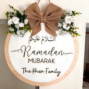 May include: A round wooden sign with a white background and a brown burlap bow. The sign says "Ramadan Mubarak" and "The Khan Family" in black lettering. The sign is decorated with white flowers.