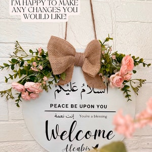 May include: A white round wooden sign with the Arabic phrase "السلامُ عَلَيْكُمْ" which translates to "Peace be upon you" in English. The sign also says "You're a blessing" in English and Arabic. The sign also says "Welcome" and "Alcabir" in black lettering. The sign is decorated with a burlap bow and pink flowers.