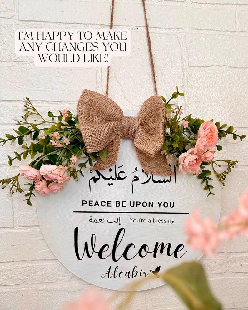 May include: A white round wooden sign with the Arabic phrase "السلامُ عَلَيْكُمْ" which translates to "Peace be upon you" in English. The sign also says "You're a blessing" in English and Arabic. The sign also says "Welcome" and "Alcabir" in black lettering. The sign is decorated with a burlap bow and pink flowers.