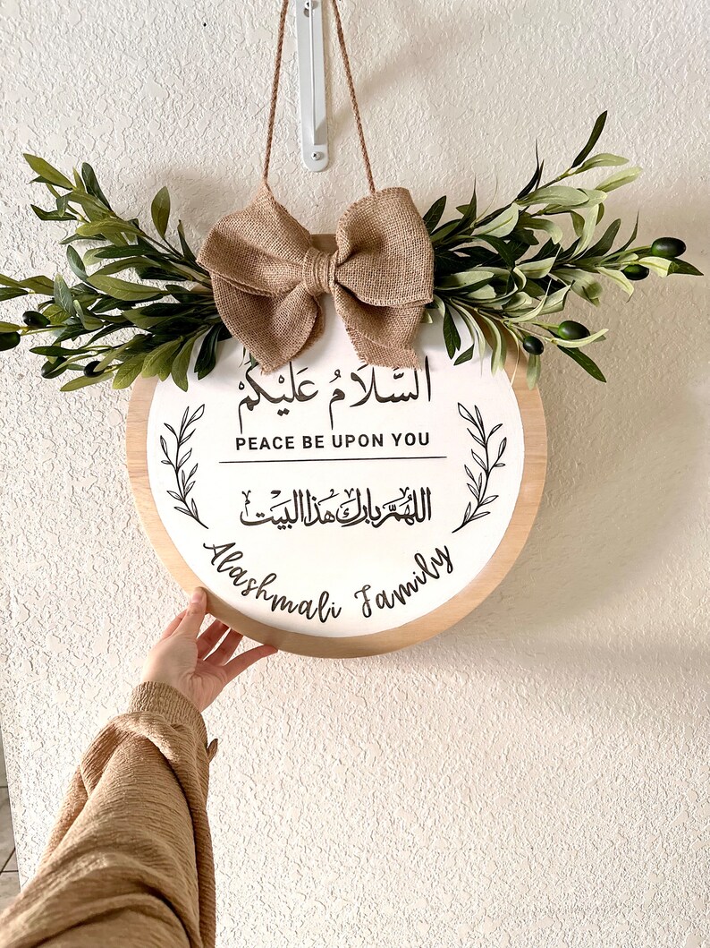 May include: A round wooden sign with the Arabic phrase "Al Salam Alaykum" which translates to "Peace be upon you" in English. The sign also includes the phrase "Allah Yarham Alladhina Mati" in Arabic, which translates to "May God have mercy on the deceased" in English. The sign is decorated with a burlap bow and olive branches. The text "Alashmali Family" is written below the Arabic phrases.