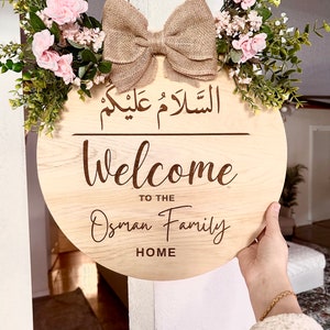 May include: A round wooden sign with the text "Welcome to the Osman Family Home" and the Arabic phrase "Al Salam Alaykum" in a decorative font. The sign is decorated with a burlap bow and a floral arrangement of pink flowers and green leaves.