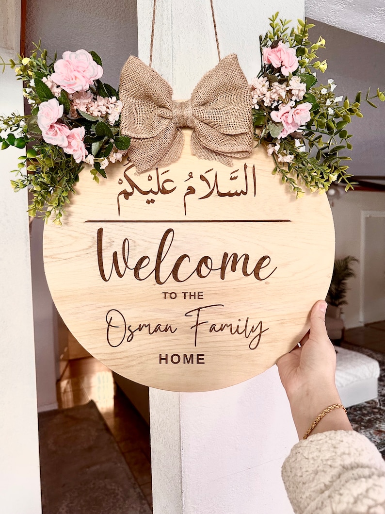 May include: A round wooden sign with the text "Welcome to the Osman Family Home" and the Arabic phrase "Al Salam Alaykum" in a decorative font. The sign is decorated with a burlap bow and a floral arrangement of pink flowers and green leaves.