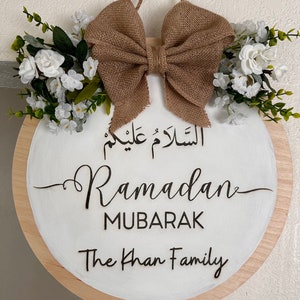 May include: A round wooden sign with a white background and a brown burlap bow. The sign says "Ramadan Mubarak" in Arabic and English, and "The Khan Family".