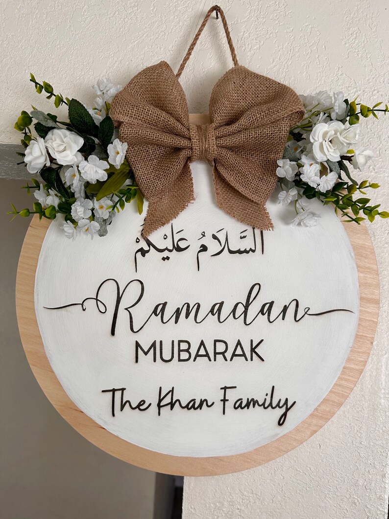 May include: A round wooden sign with a white background and a brown burlap bow. The sign says "Ramadan Mubarak" in Arabic and English, and "The Khan Family".