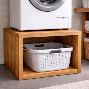 Puede incluir: Una lavadora blanca se encuentra sobre un mueble de madera con una cesta de lavandería blanca en su interior. El mueble es de madera de color marrón claro y está sobre un suelo gris. La cesta tiene un diseño perforado.