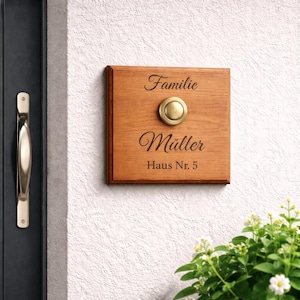 Könnte beinhalten: Ein hölzernes Klingelschild mit dem Familiennamen „Müller“ und der Adresse „Haus Nr. 5“ in schwarzer Schrift. Das Schild hat einen goldfarbenen Klingelknopf in der Mitte. Das Schild ist an einer strukturierten weißen Wand angebracht.