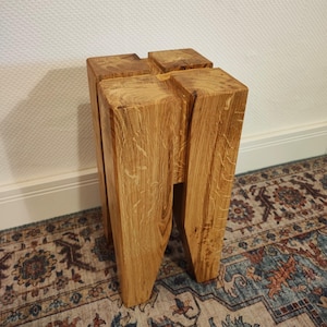 Puede incluir: Un taburete de madera con una tapa cuadrada y cuatro patas. El taburete está hecho de madera marrón claro con un acabado natural.
