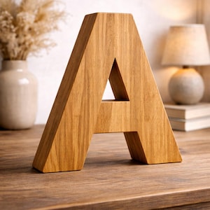May include: A wooden letter 'A' in a light brown hue, with visible wood grain, sits on a wooden table. The background features a vase with dried flowers, a lamp, and books, creating a warm, decorative setting.