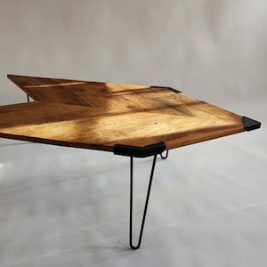 Può includere: Un tavolino da caffè in legno dal design angolare unico. Il piano è in legno marrone chiaro con un motivo a venature visibile. Presenta angoli in metallo nero e gambe a forcina nere. Il tavolo è lungo circa 122 cm.