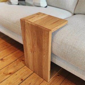 Könnte beinhalten: Ein Beistelltisch aus Holz mit natürlichem Finish. Der Tisch ist so konzipiert, dass er über die Armlehne eines Sofas geschoben werden kann. Der Tisch ist aus hellbraunem Holz und hat eine rechteckige Form. Der Tisch ist 30 cm breit, 46 cm lang und 61 cm hoch.