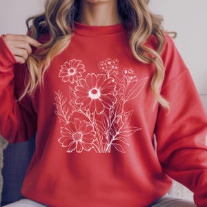 May include: A red sweatshirt featuring a white floral design. The graphic includes detailed outlines of daisies and other flowers. The sweatshirt has a crew neck and long sleeves, perfect for casual wear.