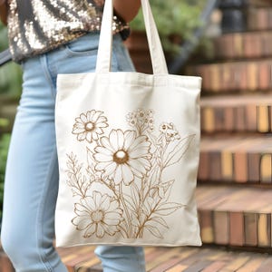 May include: Cream-colored tote bag featuring a detailed brown floral design. The illustration includes large daisies and smaller wildflowers, with stems and leaves. The bag has two long handles and is held by a person wearing blue jeans.
