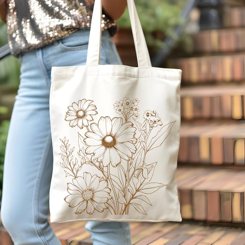 May include: Cream-colored tote bag featuring a detailed brown floral design. The illustration includes large daisies and smaller wildflowers, with stems and leaves. The bag has two long handles and is held by a person wearing blue jeans.