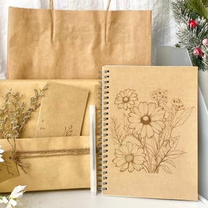 May include: A spiral-bound notebook with a floral design, a white pen, and a brown paper bag. The notebook features a detailed illustration of daisies and other flowers. The items are arranged on a white surface, suggesting a gift set.