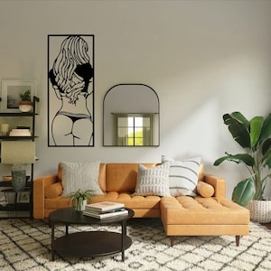 Könnte beinhalten: Ein schwarzes Wandbild aus Metall, das die Silhouette einer Frau in einem Bikini darstellt. Die Kunst ist an einer weißen Wand über einem braunen Ledersofa mit Kissen angebracht. Ein runder Couchtisch mit Büchern steht vor dem Sofa. Ein Spiegel mit einem gewölbten Oberteil hängt an der Wand rechts neben dem Kunstwerk. Eine Topfpflanze mit grünen Blättern steht in der Ecke des Raumes.