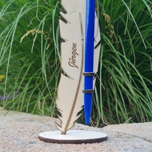 May include: A wooden feather pen holder with the name "Jürgen" engraved on it. The holder is made of 4 mm plywood and is designed to hold a pen. The holder is shown with a blue pen inserted into it.