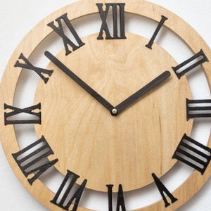 Könnte beinhalten: Eine runde Holzuhr mit schwarzen römischen Ziffern am Rand. Das Zifferblatt der Uhr ist aus hellem Holz mit schwarzen Zeigern.