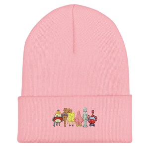 May include: A light pink beanie featuring an embroidered design of cartoon characters. The characters are in various colors, including yellow, red, and blue. The beanie is made of a soft, knitted material.