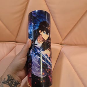 May include: A cylindrical tumbler featuring a vibrant anime-style illustration of a character with dark hair and glowing blue eyes, surrounded by dynamic blue energy effects against a dark, atmospheric background.
