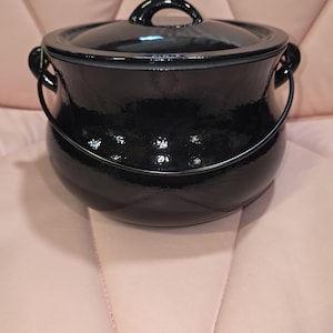Puede incluir: Una olla de cerámica negra brillante con tapa a juego y un asa de metal curvada. La olla presenta un cuerpo redondeado con dos asas laterales y un asa central en la tapa, mostrando un acabado liso y reflectante en toda la pieza.