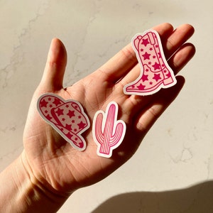 Puede incluir: Se muestran tres pegatinas rosas con contornos blancos. Las pegatinas incluyen un sombrero de vaquero con diseños de estrellas, un cactus y una bota de vaquero con diseños de estrellas. Las pegatinas se sostienen en una mano.