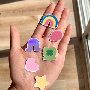 May include: A hand holding a collection of colorful stickers. The stickers include a rainbow, a balloon, a four-leaf clover, a heart, a star, and a holographic shape. The stickers are on a wooden surface.