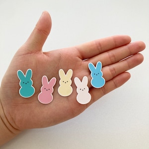 Puede incluir: Cinco adornos con forma de conejito en tonos pastel, azul, rosa, amarillo y blanco, cada uno con un contorno blanco y tres pequeños puntos negros para los ojos. Los adornos están dispuestos sobre una superficie clara.