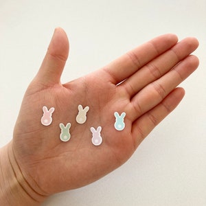 Puede incluir: Una mano sostiene seis pegatinas pastel con forma de conejito. Las pegatinas son de color rosa, verde, amarillo y azul, cada una con una cola blanca en forma de estrella. Son pequeñas y delicadas, perfectas para manualidades o decoración.