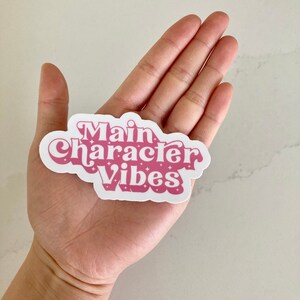 May include: A white sticker with the text "Main Character Vibes" in a retro pink font. The sticker has a cloud-like shape and is held in a hand.