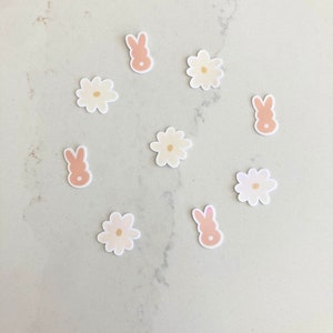 Puede incluir: Una colección de pegatinas con temática primaveral. Las pegatinas incluyen formas de conejos rosas claros y flores blancas con un centro amarillo claro. Las pegatinas están dispersas sobre una superficie gris claro.