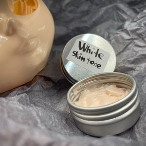May include: A small, silver tin with a white label that reads "White Skin Tone". The tin is open and contains a pink, creamy substance.