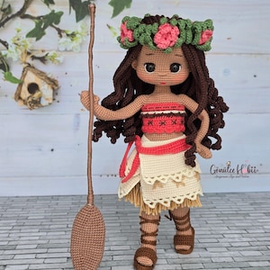 Puede incluir: Muñeca de ganchillo hecha a mano que representa a un personaje con piel morena, pelo largo castaño y una corona floral. La muñeca viste un top rojo y blanco, una falda crema y sandalias marrones. Sostiene un bastón marrón.