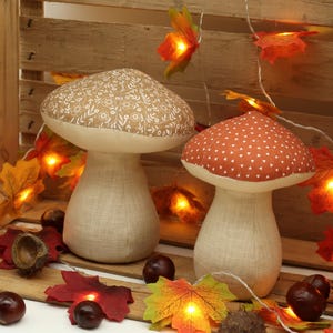 May include: Two fabric mushroom decorations with a brown and white patterned design and a red and white patterned design. The mushrooms are sitting on a wooden surface with a string of lights with small maple leaf-shaped bulbs.