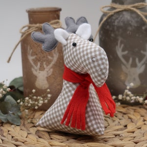 May include: A gray and white plaid moose with a red scarf. The moose is sitting on a woven wicker placemat with a small bouquet of white flowers and a red ornament.
