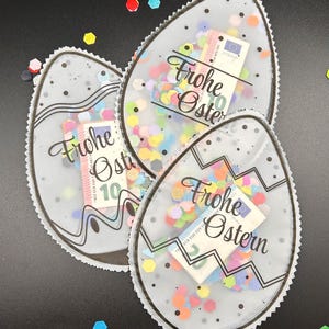 Könnte beinhalten: Drei transparente, eiförmige Geschenkbeutel gefüllt mit bunten Konfetti und einem Euro-Schein. Jeder Beutel hat eine schwarze Umrandung und die Worte "Frohe Ostern" in Schreibschrift. Die Beutel liegen auf einer schwarzen Oberfläche mit verstreuten Konfetti.