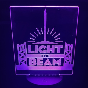 May include: A purple LED light with the words "LIGHT THE BEAM" etched into the acrylic. The design includes a stylized beam of light and architectural elements. The base is black and round.