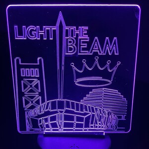 May include: A purple LED night light with the words "LIGHT THE BEAM" and a graphic of a bridge, a tower, a crown, and a building. The night light has a black base with a power symbol.