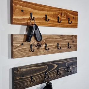 May include: Three wooden key racks with black metal hooks mounted on a white wall. The top rack is stained a light brown, the middle rack is stained a medium brown, and the bottom rack is stained a dark brown.