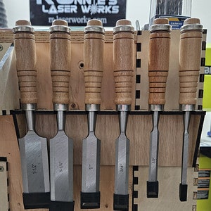Könnte beinhalten: Ein Satz von sechs Holzgriff-Meißeln mit Metallklingen, die an einem Holzgestell hängen. Die Meißel sind mit ihren Größen beschriftet: 1-1/2", 1-1/4", 1", 3/4", und 1/2".