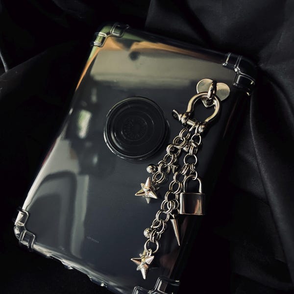 Puede incluir: Una funda transparente para teléfono con protectores de esquina plateados y un agarre circular negro. Una cadena plateada con dijes de estrella y candado cuelga de la funda, creando una estética gótica.