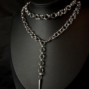 May include: A silver-toned necklace with a layered design. The necklace features a chain with large, rounded links and a vertical drop with a series of smaller rings and a pointed pendant. The jewelry is displayed on a black mannequin.