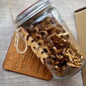Può includere: Un barattolo di vetro pieno di tessere di lettere in legno, con la parola "F**K" scritta in tessere più grandi. Il barattolo è appoggiato su un sottobicchiere di legno con la parola "F**K" incisa su di esso più volte.