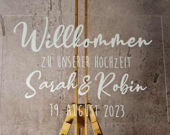 Op de afbeelding: Een helder acryl bord met witte letters met de tekst "Willkommen zu unserer Hochzeit Sarah & Robin 19. August 2023".